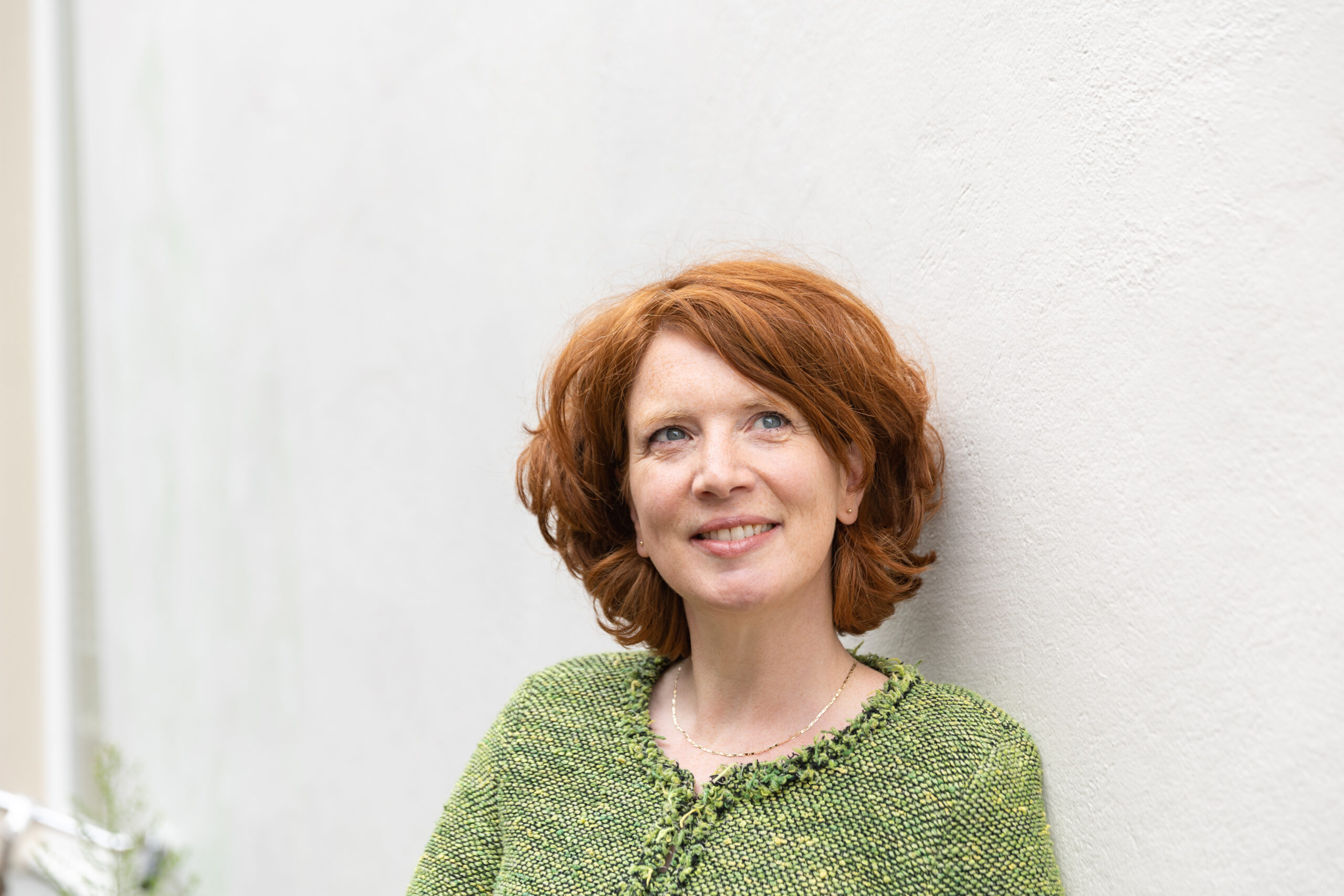 Vrouw met rood haar en een groene trui die leunt tegen een grijze muur. Ze kijkt iets schuin omhoog.